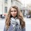 Headshot of charming Caucasian young woman with light-brown wavy hair dressed in stripped coat and big grey scarf staring at camera with lazy smile standing on European city background